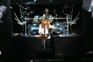 Dave Mustaine, de camisa branca e longos cabelos ruivos cacheados, toca sua guitarra flying v em show do Megadeth