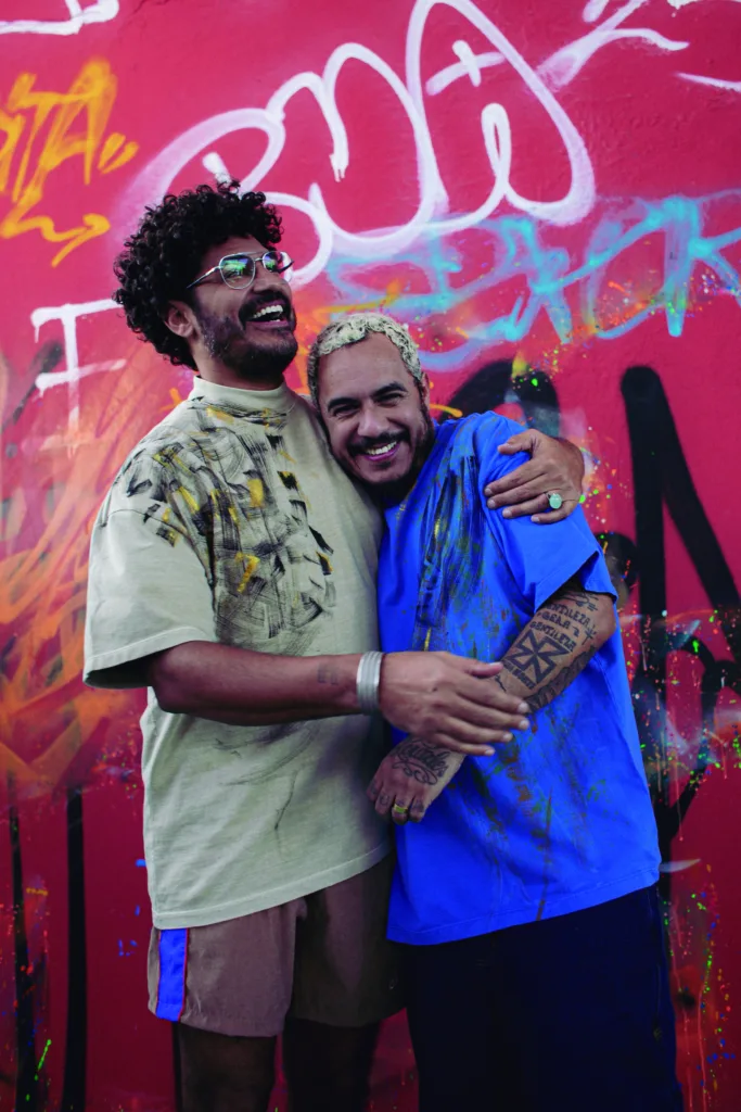 Criolo abraça Marcelo D2, os dois estão na frente de uma parede vermelha com pixos. Eles usam camisetas também pixadas.