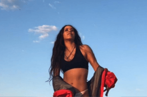 A cantora Flora Matos posa de top preto, calcinha clara e casaco sobre os ombros na frente do céu azul