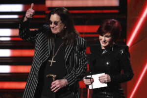 O cantor Ozzy Osbourne, todo de preto, faz sinal de joinha no palco do Grammy, enquanto sua mulher, Sharon, segura o envelope com o nome de um dos vencedores da cerimônia de 2020