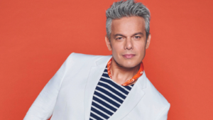 Otaviano Costa, homem branco de cabelos grisalhos, com camiseta listrada e blazer branco sobre fundo vermelho